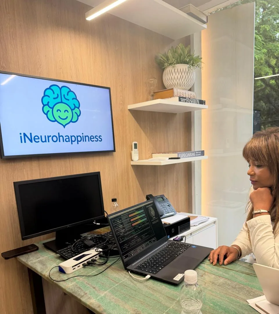 Anna Oliver sentada à mesa de trabalho, diante de uma tela com o logo do iNeurohappiness